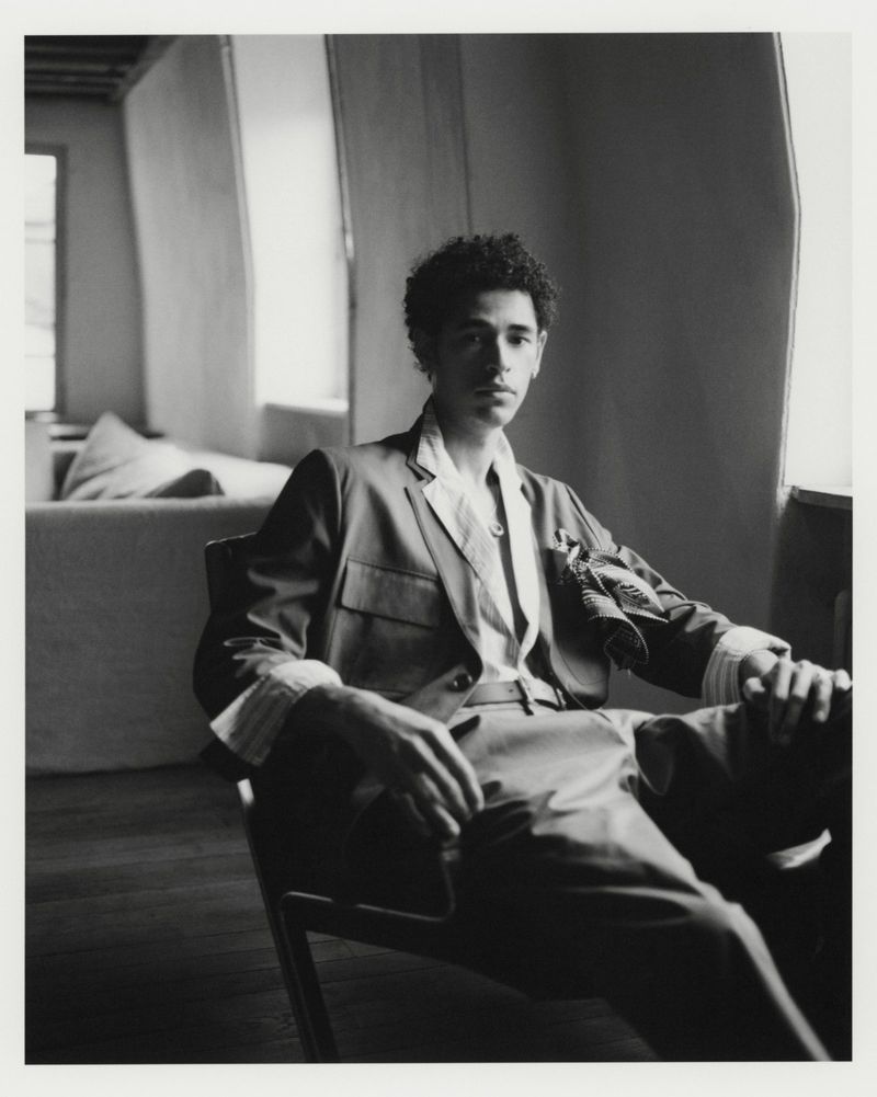 A man in a tailored jacket with cuffed sleeves and trousers sits in an armchair beside a window in a black and white portrait.