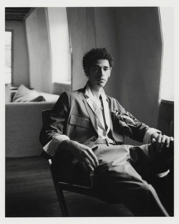 A man in a tailored jacket with cuffed sleeves and trousers sits in an armchair beside a window in a black and white portrait.