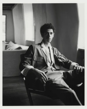 A man in a tailored jacket with cuffed sleeves and trousers sits in an armchair beside a window in a black and white portrait.