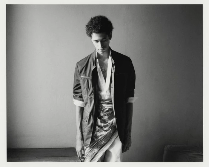 Black and white portrait of a young man wearing a dark jacket, unbuttoned white shirt, and patterned silk scarf tied at the waist.