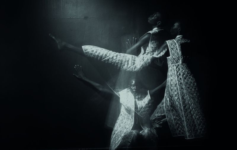 Monochrome portrait of a Black woman in motion, wearing an ornate dress. She is captured in multiple transparent overlapping images.