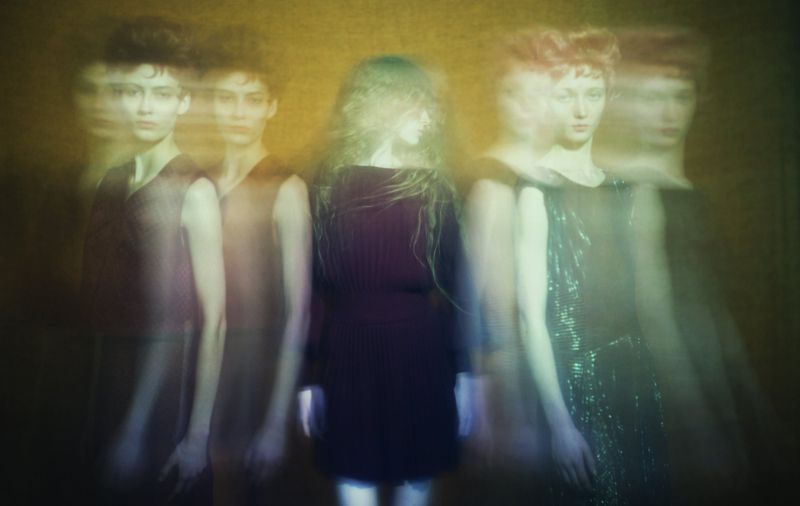 Five young adult women in dresses stand in a blurred, layered fashion photo with a retro aesthetic and warm yellow and green lighting.