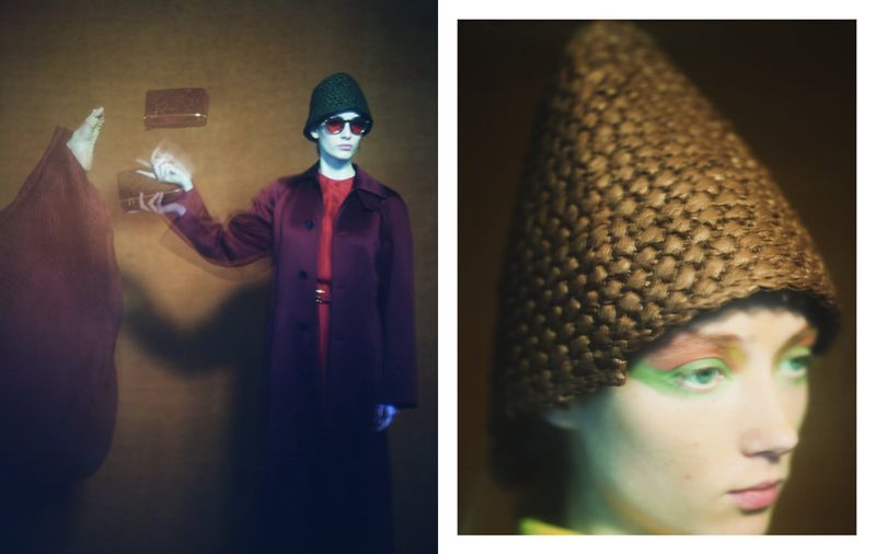 A woman in a maroon coat and green hat holds a purse in a studio with warm lighting and a blurred figure, creating a vintage, fashion-forward aesthetic.