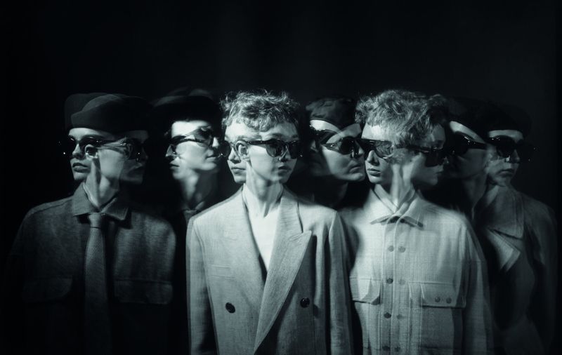 Monochrome image of a man in a jacket and sunglasses with a motion blur effect, creating a multiple exposure look, set against a dark background.