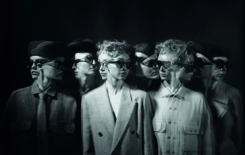 Monochrome image of a man in a jacket and sunglasses with a motion blur effect, creating a multiple exposure look, set against a dark background.