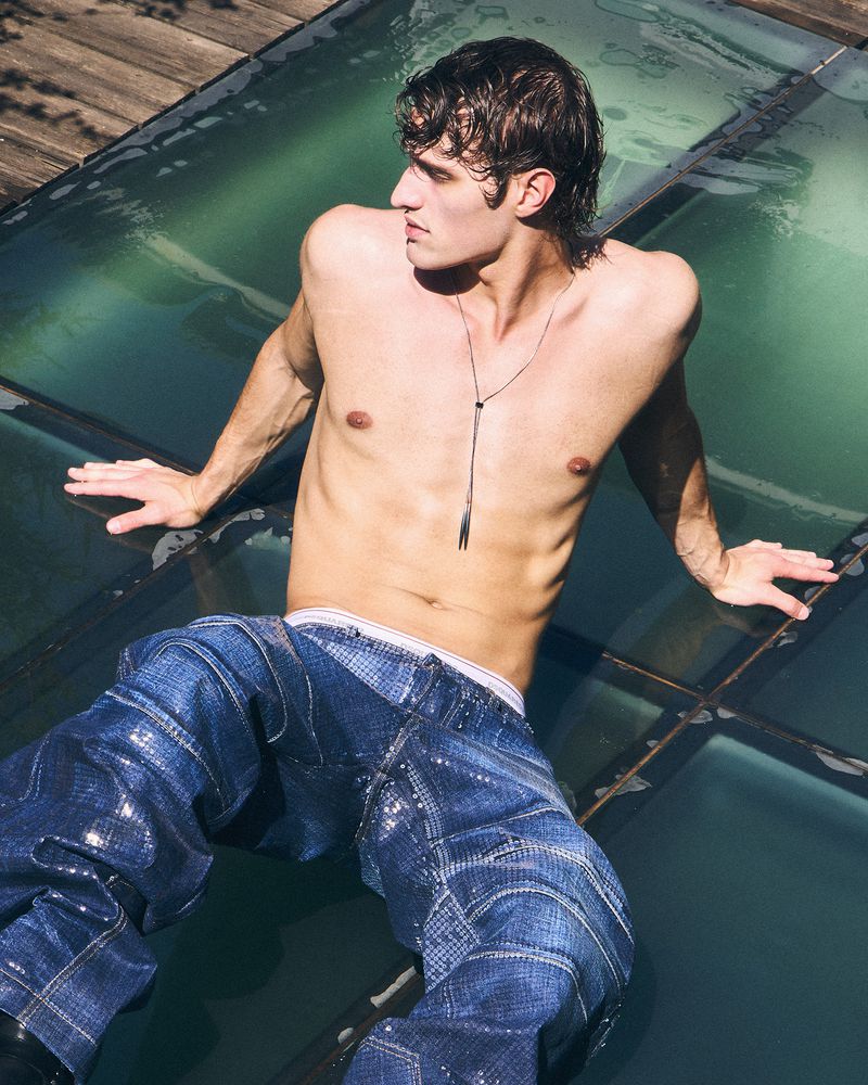 Shirtless male model wearing textured blue jeans reclining outdoors on glass panels in natural sunlight.