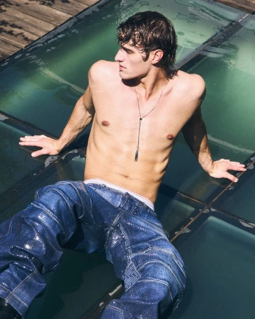 Shirtless male model wearing textured blue jeans reclining outdoors on glass panels in natural sunlight.