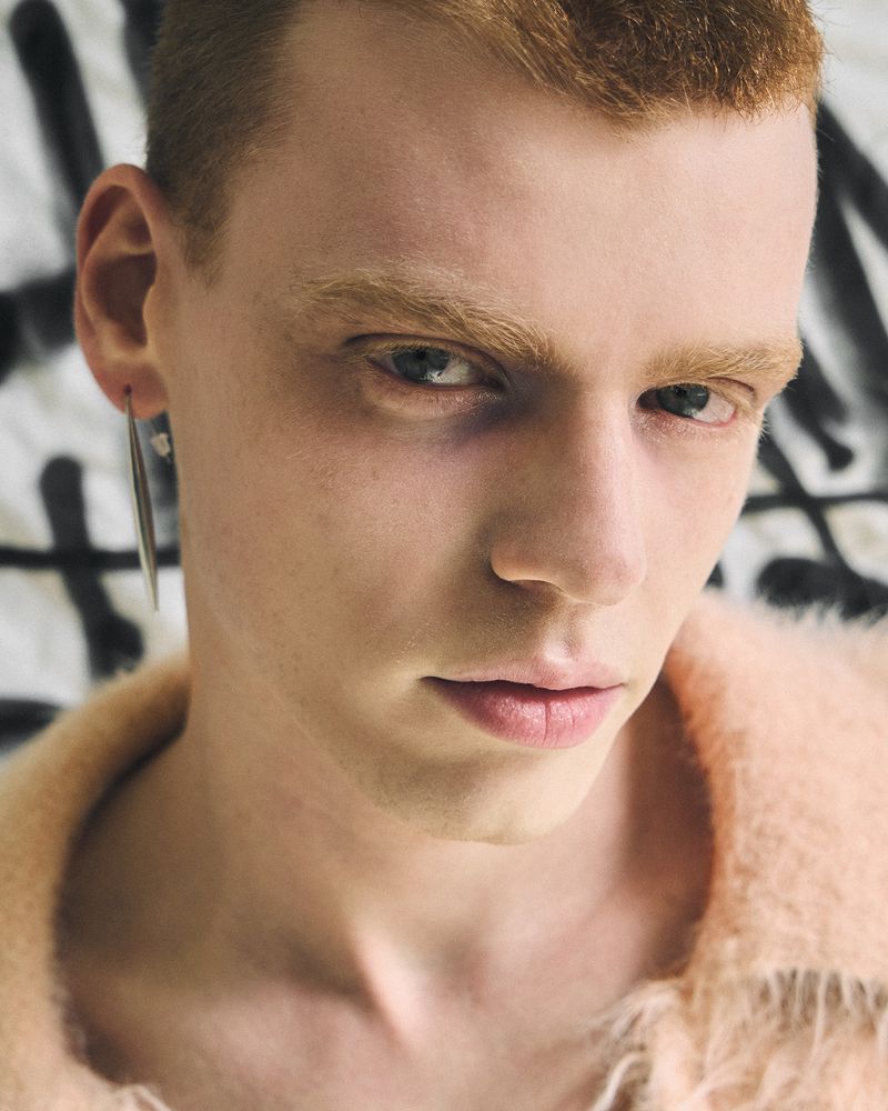Close up portrait of a young man with red hair wearing a fuzzy pink sweater and a long silver earring against a patterned background