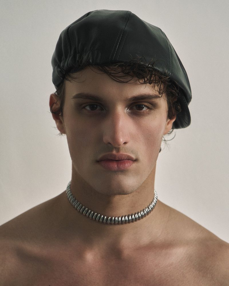 Close-up portrait of a man wearing a black leather beret and a silver coiled choker necklace against a neutral backdrop.