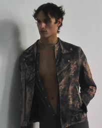 A male model in an open distressed bleach-patterned leather biker jacket and silver choker necklace posing against a light background.