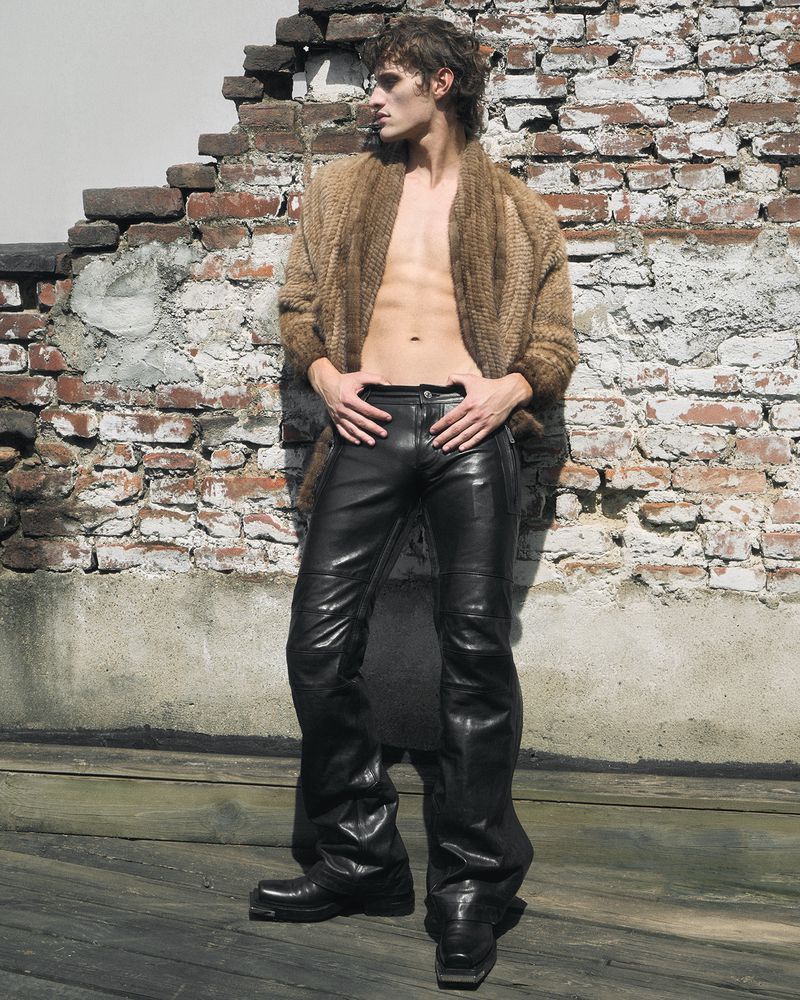 A man posing in an open brown cardigan and black leather pants against a weathered brick wall.