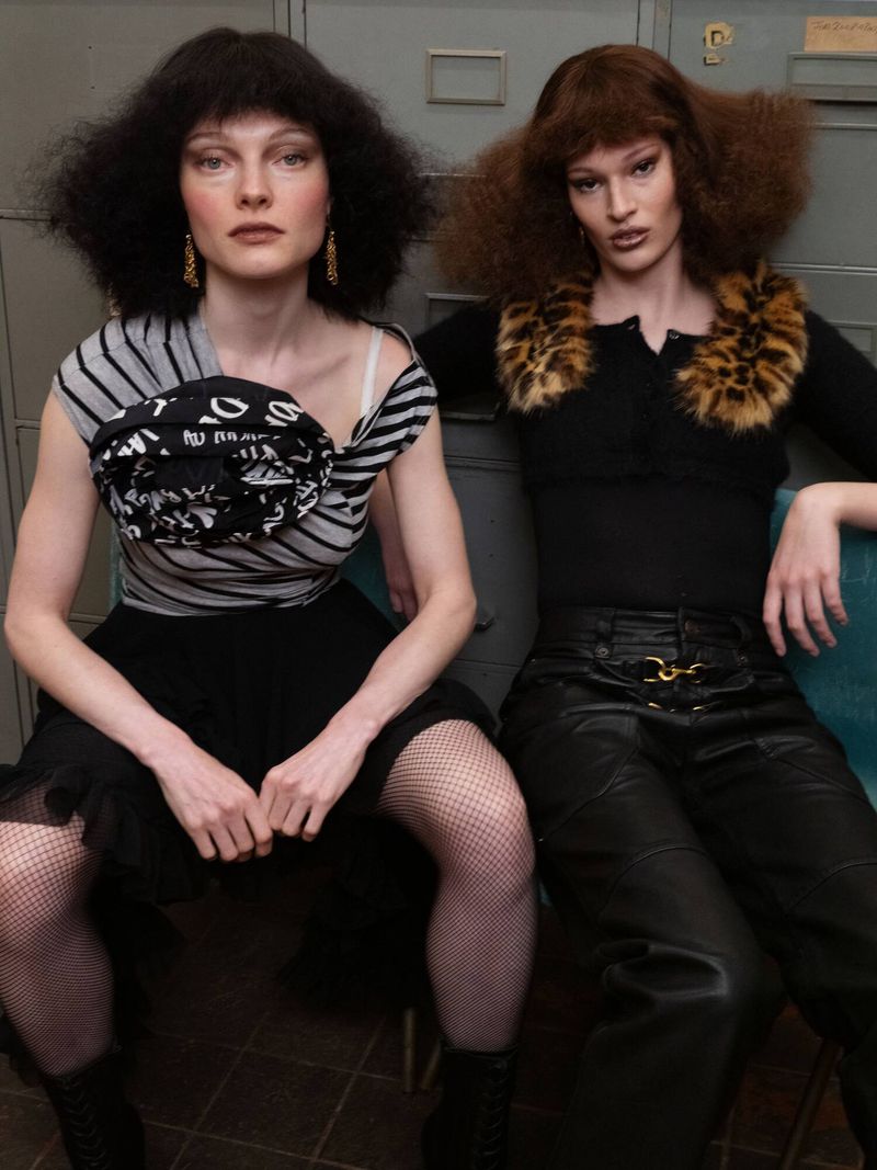 Two fashion models with voluminous retro hairstyles sitting against grey vintage office cabinets wearing textured black outfits.
