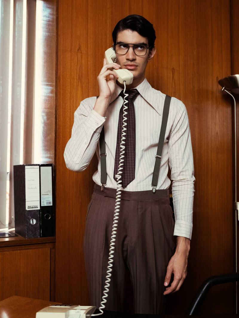 Man in retro office attire wearing glasses and suspenders while talking on a landline telephone in a wood-paneled room.