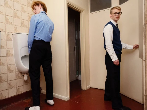 Two young men in school-uniform style menswear posing inside a retro tiled restroom with a urinal and cubicle doors.