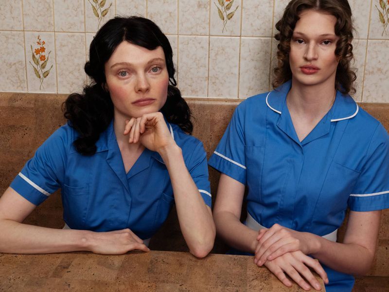 Two women in matching blue short-sleeved uniforms seated at a table against a floral-tiled wall.