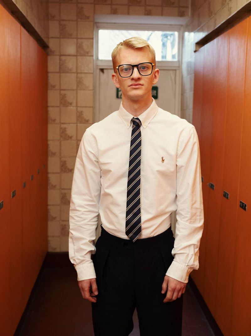 A blonde young man wearing black-rimmed glasses, a white dress shirt, striped tie, and black trousers stands in an orange locker hallway.