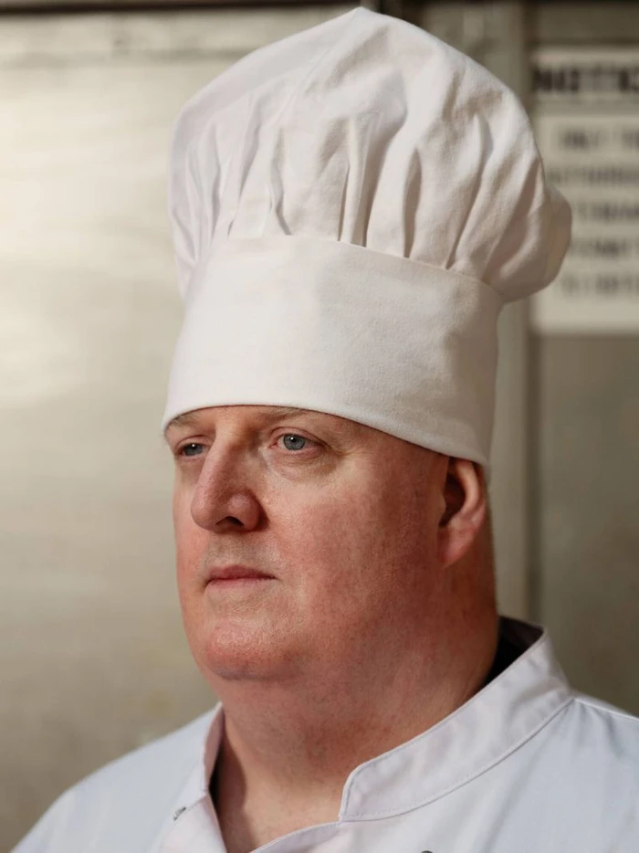 Close-up portrait of a man wearing a tall white chef hat and chef uniform against a stainless steel background.