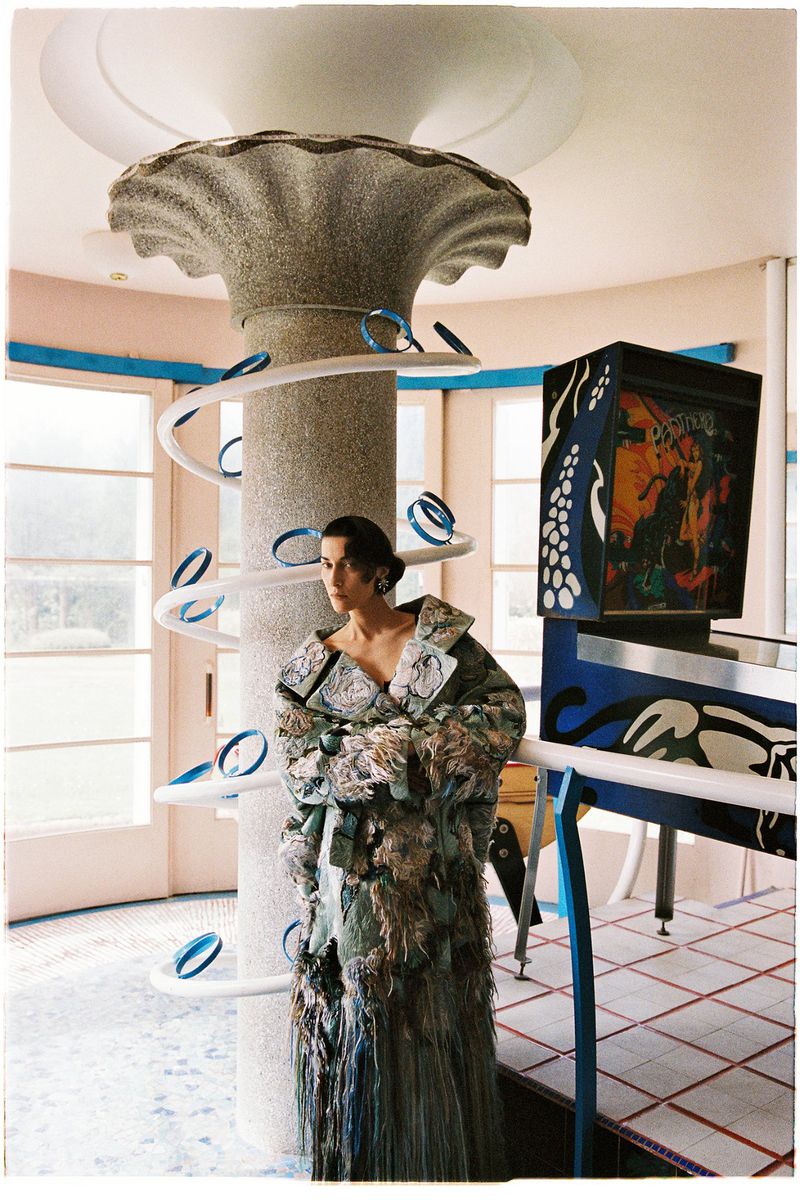 Fashion model wearing a textured fringe coat standing by a spiral column next to a vintage pinball machine.