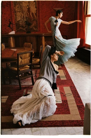 Two women in elegant dresses pose inside an ornate red room, one reclining on a geometric rug and one standing near a window.