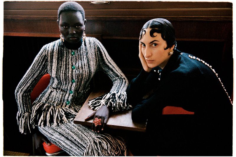 Two female models seated at a wooden table in high fashion knitwear, one in fringed tweed and one in sleek black attire.