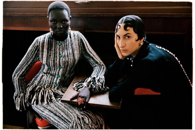 Two female models seated at a wooden table in high fashion knitwear, one in fringed tweed and one in sleek black attire.
