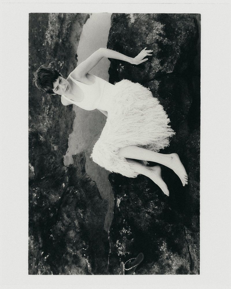 Black and white photograph of a woman in a white fringe skirt reclining across dark coastal rocks.