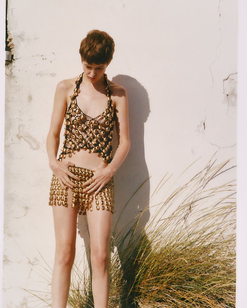 Model Lando van der Schee posing in a metallic gold embellished top and skirt set against a sunlit white wall with tall grasses.