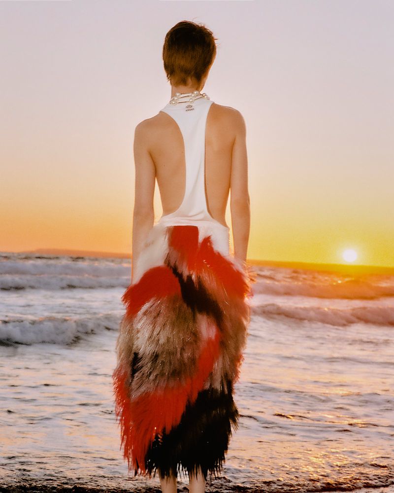 Model standing on a wet sandy beach at sunset seen from behind wearing an open-back white top and colorful feathered skirt.