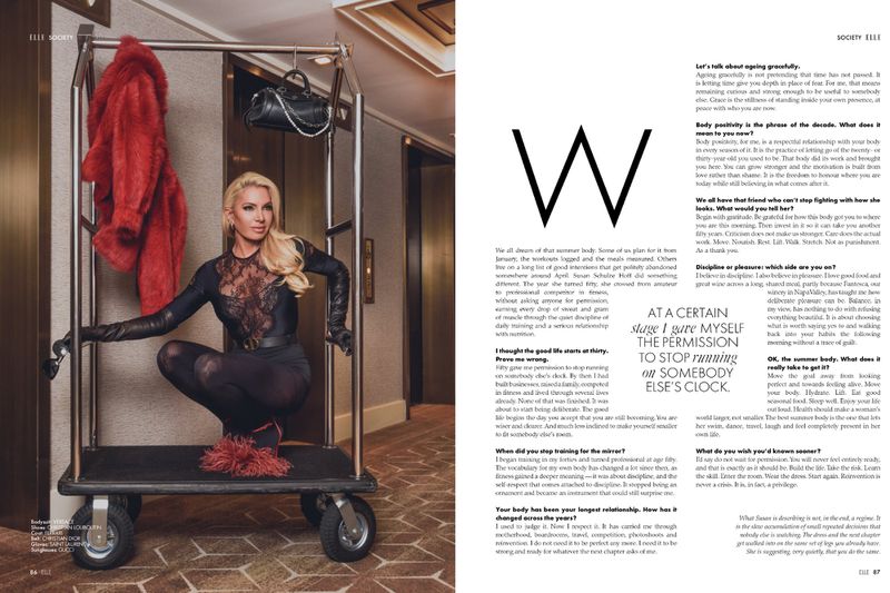 Blonde woman crouching on a luggage cart wearing a black lace bodysuit and gloves, with a red coat hanging beside her.