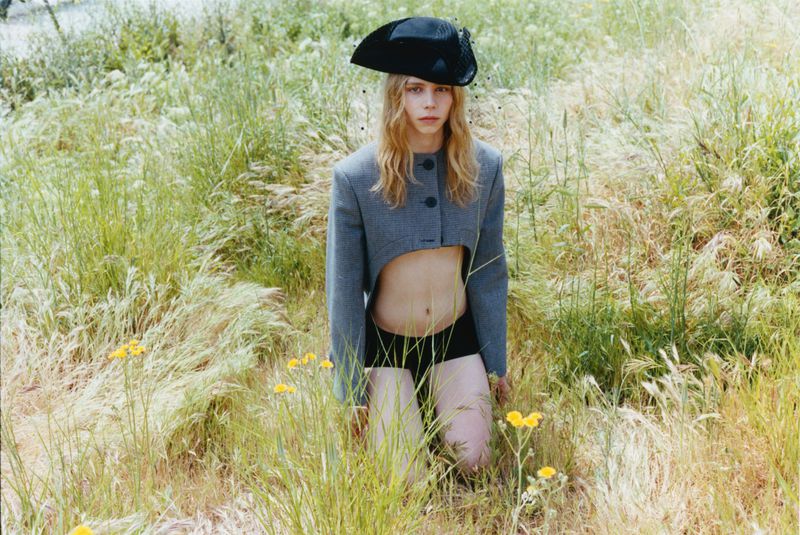 Model Rafe Crane-Robinson kneeling in tall grass wearing a cropped grey blazer, black briefs, and a black sculptural hat with a veil.