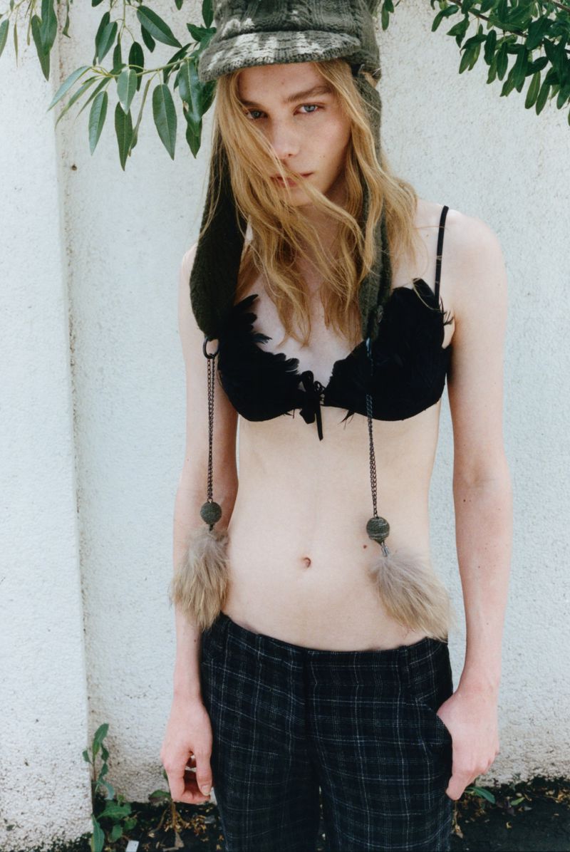 Model Rafe Crane-Robinson wearing a trapper hat with pom-poms, black bra top, and plaid pants against a pale textured wall.