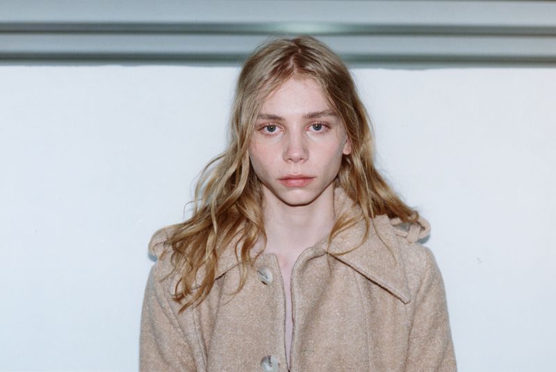 Portrait of a young model with long blonde hair wearing a camel buttoned coat against a plain white wall