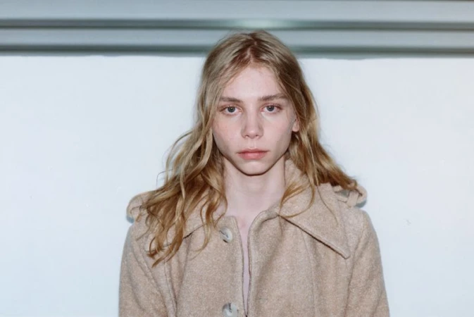 Portrait of a young model with long blonde hair wearing a camel buttoned coat against a plain white wall