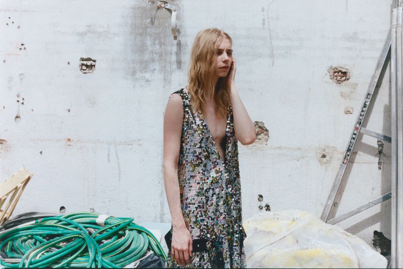 Model Rafe Crane-Robinson in a shimmering sequined sleeveless dress beside coiled green hoses and a ladder.