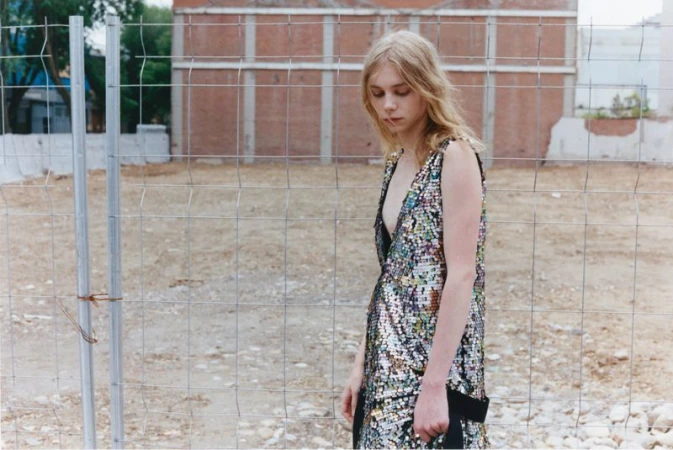 Model wearing a sequin dress standing outdoors behind a wire mesh fence with a brick building in the background.