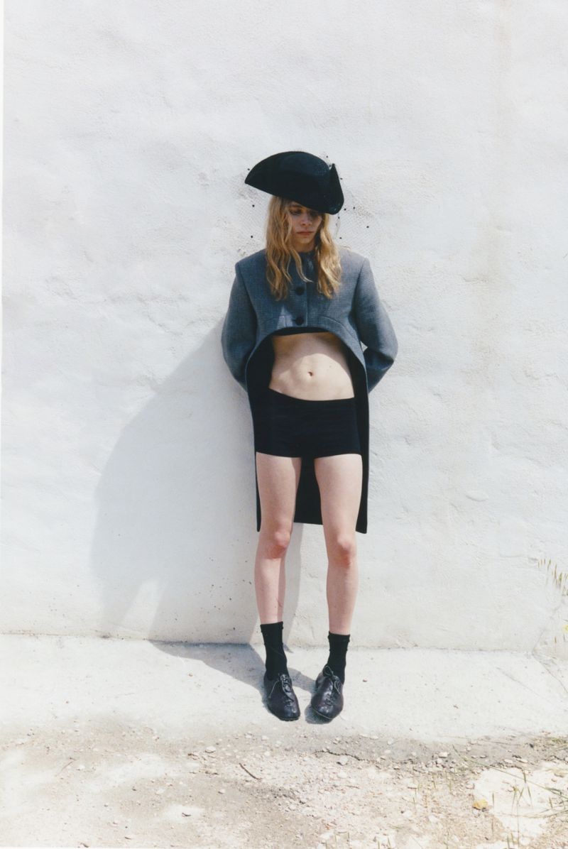 Model wearing a cropped grey coat, tricorn hat, and black shorts standing against a bright whitewashed wall.