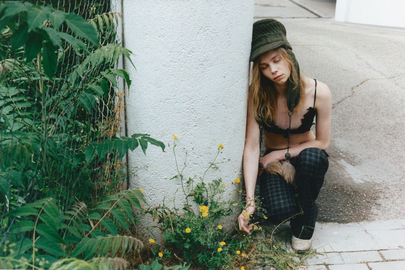 Model Rafe Crane-Robinson crouching beside a white pillar wearing an olive knit trapper hat, black bralette, and dark plaid trousers.