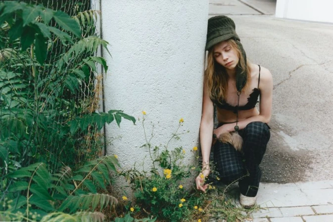 Model Rafe Crane-Robinson crouching beside a white pillar wearing an olive knit trapper hat, black bralette, and dark plaid trousers.