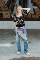Model posing outdoors in a black reindeer knit top, blue jeans, and a grey sweater tied around the waist in front of cardboard boxes.