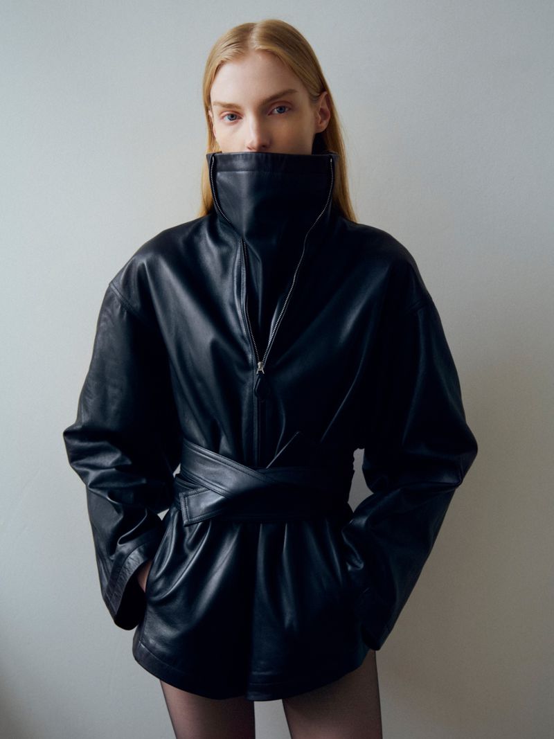 Model wearing a high-collared black leather jacket with a belted waist against a neutral grey studio background.