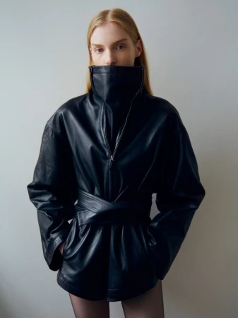 Model wearing a high-collared black leather jacket with a belted waist against a neutral grey studio background.