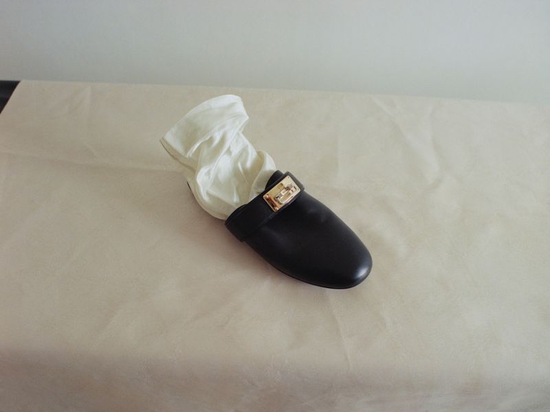 A single black leather mule shoe with a gold buckle and white cloth inside, resting on a neutral beige draped surface.