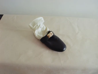 A single black leather mule shoe with a gold buckle and white cloth inside, resting on a neutral beige draped surface.