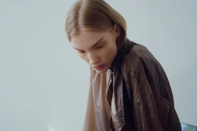 Model Alexandra Liv looking downward while wearing a brown collared jacket against a neutral studio backdrop.