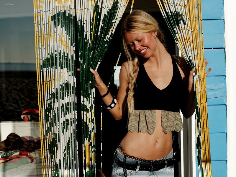 Woman with blonde braid wearing a black cropped top and low-rise jeans smiling behind parted beaded curtains.