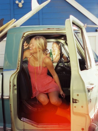 Blonde woman in a pink smocked dress sitting inside an open-door vintage pickup truck with surfboards mounted in the back.