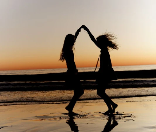 Two women silhouetted against an orange sunset dancing and holding hands on a wet sandy beach.