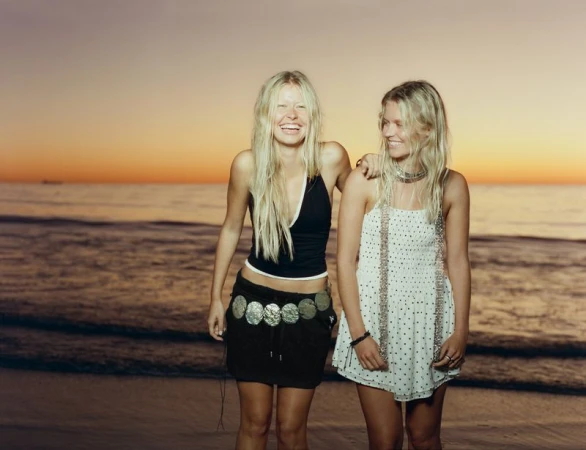 Two smiling young blond women standing on a sandy beach at sunset wearing casual summer clothing and a metallic statement belt.