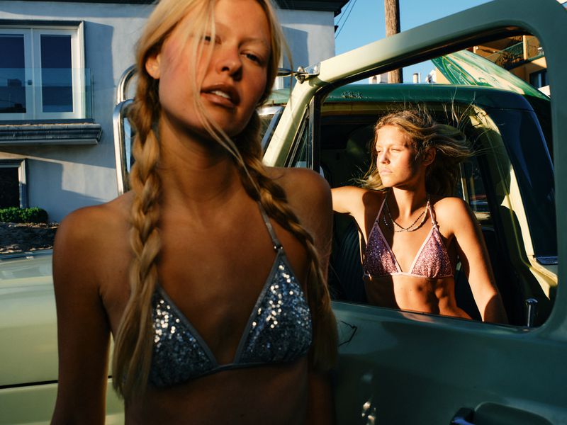 Blonde woman with braided pigtails in a silver sequin bikini top standing next to a vintage vehicle with another woman in the truck window.