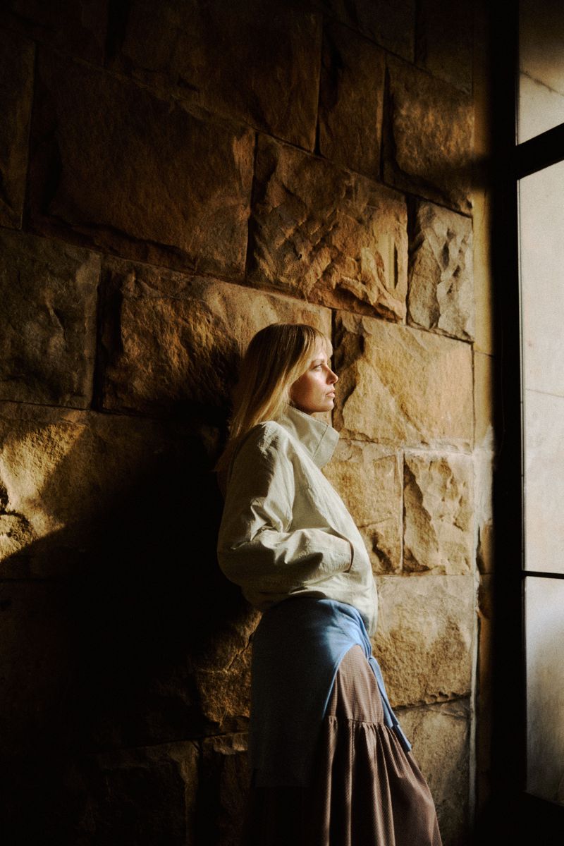 Blonde woman wearing a cream jacket and layered skirt leaning against a rustic stone wall in natural sunlight.
